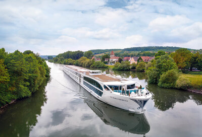 A sleek white river cruise ship moves along a narrow, calm river bordered by lush green trees, with a small village and rolling hills in the background under a partly cloudy sky.