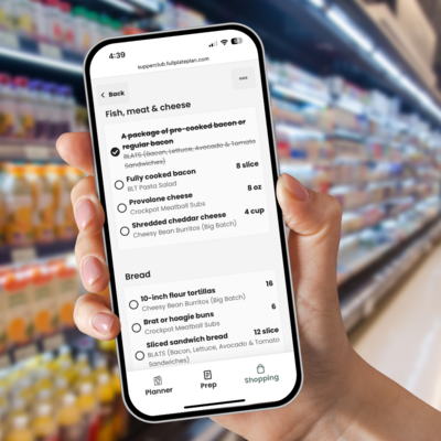Hand holding a smartphone in a grocery store aisle, displaying the Supper Club app’s digital shopping list with meal ingredients and quantities.