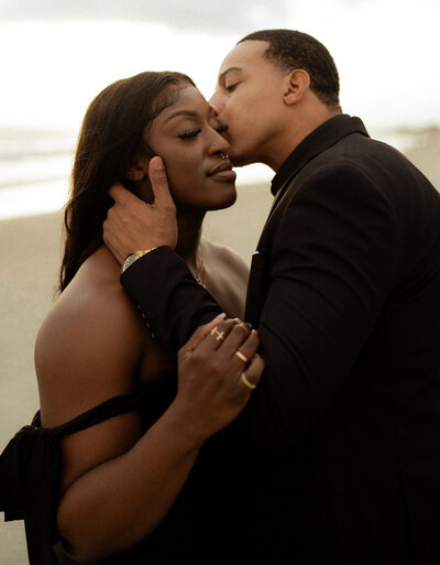 Black and white beachside photo of a man gently kissing a woman's cheek as she closes her eyes, smiling. Soft, intimate couples photography with ocean waves in the background
