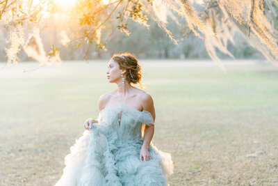 bride in a pale teal blue tulle ruffled gown standing under the spanish moss covered oak trees during golden hour at Hewitt Oaks in Bluffton SC captured by Savannah wedding photographer Magnolia West Photography
