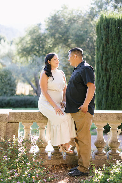 engaged couple women sites on a stone fence and male standing while the look at each other with a glow in the background
