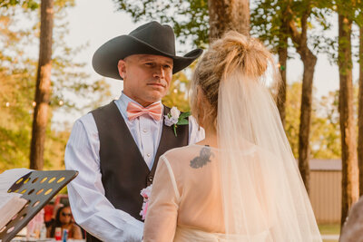Groom in a cowboy hat tearing up during the ceremony at a rustic backyard wedding in Beaumont, Texas. The bride stands in a lace gown and veil, facing him under the tall trees with soft sunlight filtering through.