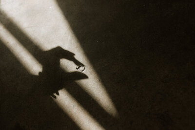 Artistic detail shot of a hand holding an engagement ring, casting a shadow in warm sunlight, photographed by Rainstorm Photo & Video in a cinematic, minimal style.