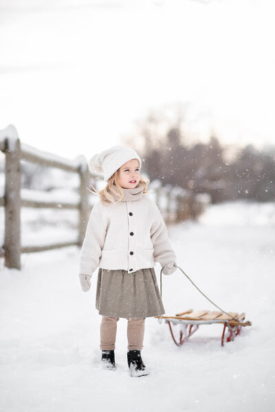 Little girl dressed in layers, pulling a sled in the snow while snows falls in the winter.  Captured by Brittany Danielle Photography