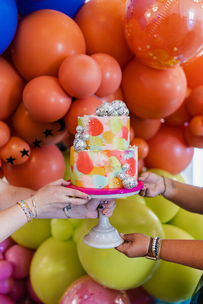 A vibrant, hand-painted two-tier cake adorned with mini disco balls and pearls is held up by four hands in front of a playful balloon backdrop in orange, yellow, and pink hues. Perfectly captures the fun and energy of a modern birthday celebration. Photo by Dani Lacey Photographs.