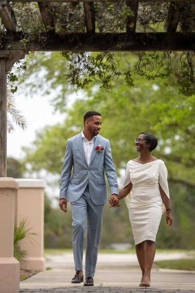 Newlywed couple holding hands and walking together under a rustic wooden pergola after their St. Augustine courthouse wedding. The groom wears a light blue suit with red boutonniere, and the bride wears an elegant white off-shoulder dress. Lush green foliage and dappled sunlight create a romantic outdoor setting.
