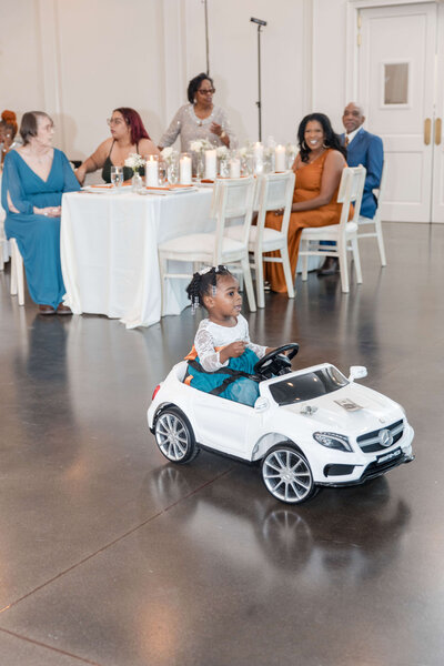 Adorable flower girl making a grand entrance in a mini luxury car at a stylish wedding reception.
