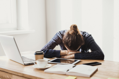 tired woman resting her head on her desk with coffee, tablet, book and phone scattered