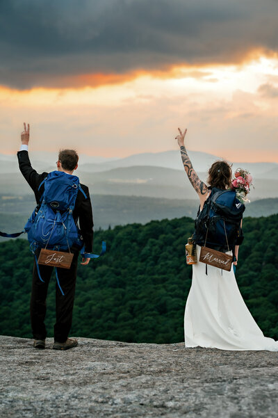 Couple throws up peace signs into the wind with a just married sign on their backpacks because they got married at the top of the mountain in the adirondacks. 