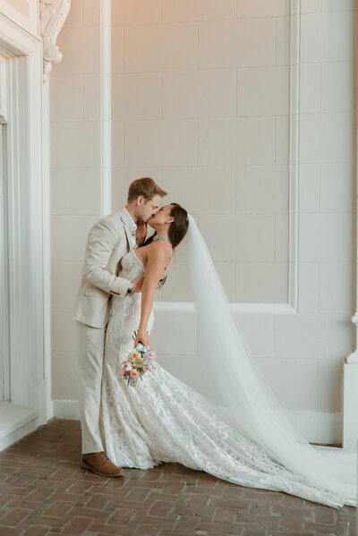 A photo of a bride and groom taken by Bailee Freeman Photography in front of the Grand Island Mansion in Walnut Grove, California. Groom is grabbing bride's waist and pulling her in tight for a kiss while her veil flows behind her. 