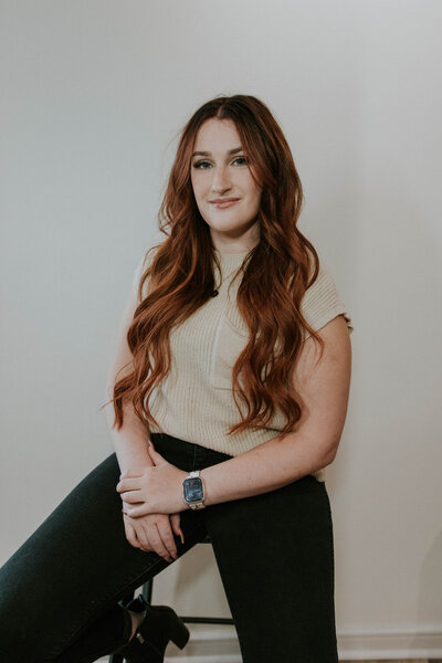 A professional headshot of Emily Brackeen, Heath Salon and Spa's HR Director. She is seated on a stool, smiling warmly with a friendly and approachable expression. The clean and minimal background highlights her professional yet inviting demeanor.