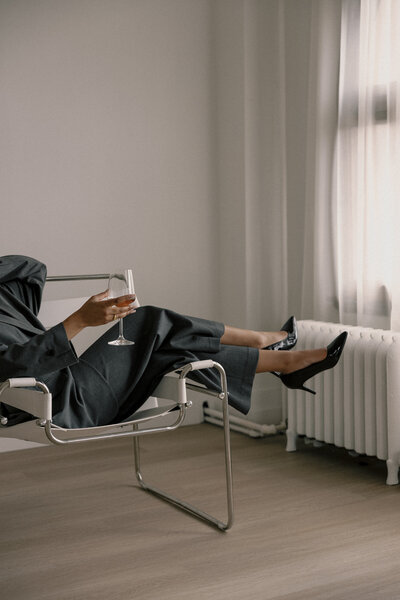 A woman in a gray suit sitting with her legs over the arm of a chair holding a glass of wine 