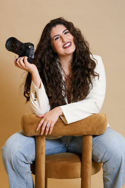 Smiling at the camera, Hiba sits backwards on a chair while holding her camera in one hand.
