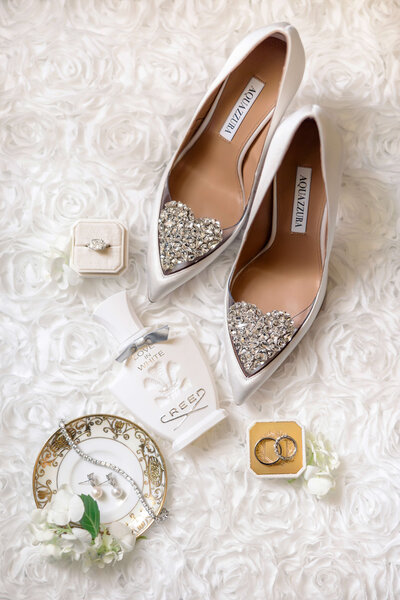 A pair of elegant silver high heels with jeweled hearts are placed on a floral textured surface. Nearby are a ring in a box, Creed perfume, a plate with earrings, and a small bouquet.