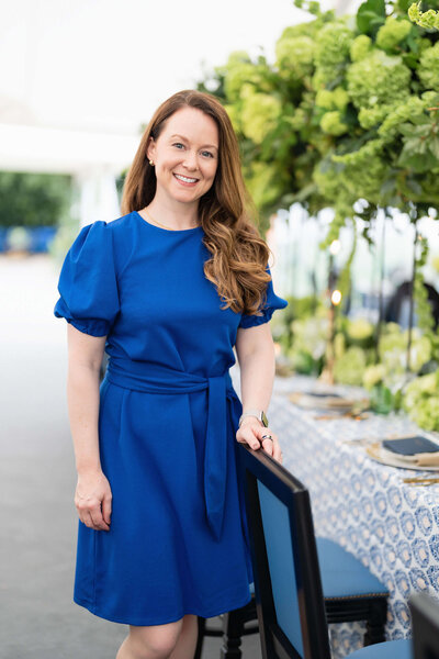 top uk wedding planner emma westacott in a blue dress standing with a blue chair at a dining table with green flowers