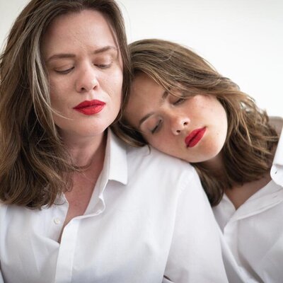 A mother and daughter sit with eyes closed, red lipstick, and oversized white button down shirts. The daughter rests her head on her mother's shoulder