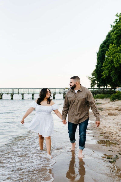 beach engagement photos with bride and groom holding hands and walking in the shore line running in the water and laughing with each other captured by Virginia wedding photographer