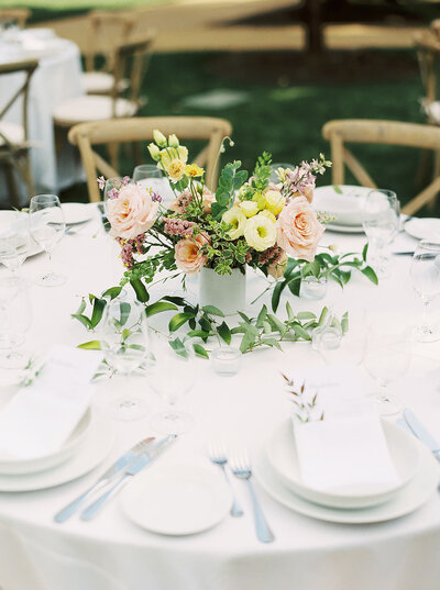 Round centerpiece of pink, orange and yellow flowers filled with greenery in a reception table outside