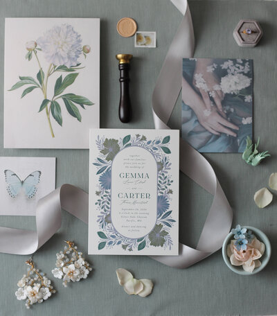 A styled collection of luxury wedding items including an invitation, wax seal and sealing wax stamp, a postage stamp, a botanical drawing of a peony, a velvet ring box with an engagement ring, beaded earrings, a vintage bluebird brooch, satin ribbon, and photographs of butterflies, hands, and textiles.