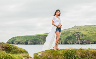 pregnancy portrait of a woman with long, brunette hair wearing a white, flowing dress while standing on a cliff overlooking the sea in Kerry