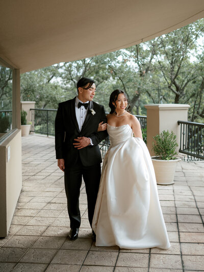 A newlywed couple walks together outdoors, smiling at each other. The bride wears an elegant strapless ball gown with a flowing train, while the groom is dressed in a classic tuxedo with a bow tie. 