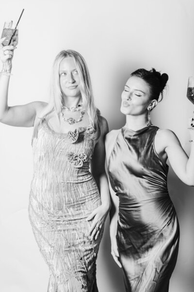 Two wedding guests in satin dresses holding cocktails and smiling in a black and white editorial-style portrait against a white backdrop.