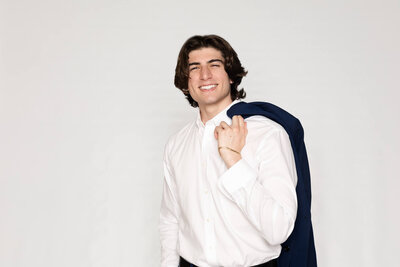 A young man with dark hair smiling at the camera in a white studio, wearing a long sleeve white dress shirt and holding his navy suit jacket over his left shoulder with his left hand.