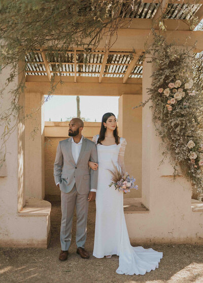Newlywed couple sands in front of camera with arms linked. They stand in front of a pueblo style arch with peach flowers