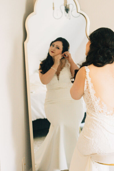 A bride admiring her reflection while adjusting her earrings in a mirror