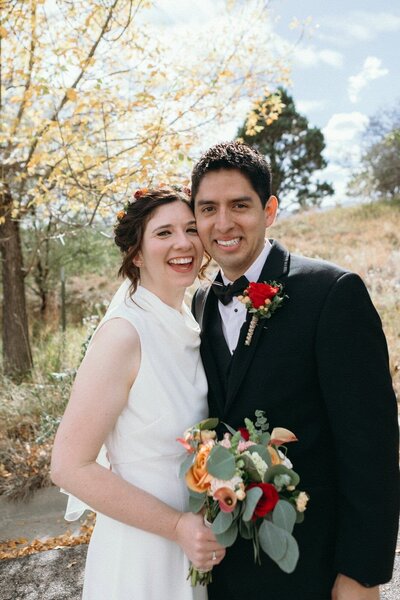 A joyful bride and groom stand cheek to cheek in front of autumn trees.