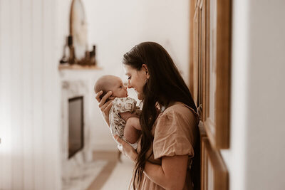 Mom cuddles newborn baby girl  nose to nose in a white hallway at home