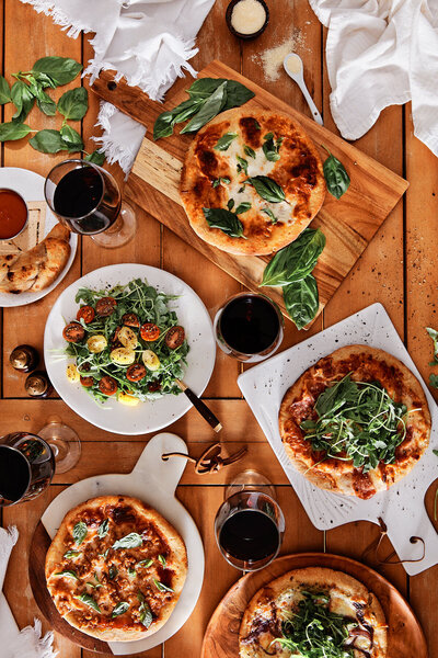 An overhead view of an assortment of gourmet pizzas and sides, beautifully arranged on a wooden table. The image, captured by Austin-based food photographer Mica McCook, highlights the variety of toppings, crispy crusts, and fresh ingredients, making it a feast for the eyes.