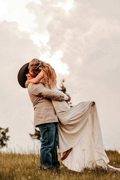 groom in a tan suit jacket and a cowboy hat  picks up his bride and she pops up her foot and leans into kiss him
