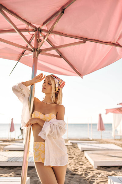 Woman in a yellow swimsuit and white cover-up standing under a pink beach umbrella on a sunny seaside.