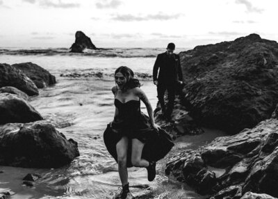 A bride in a black wedding dress runs from the incoming waves on a rocky beach as her groom steadily follows during their adventure elopement 
