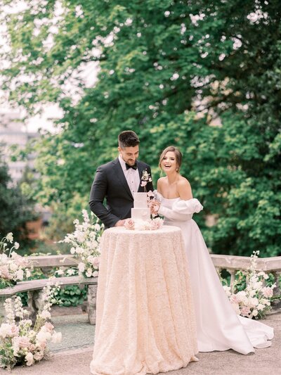 A couple stands by a round table cutting a cake outdoors, both dressed in formal wedding attire, surrounded by greenery and floral decorations. Their celebration, meticulously crafted by a top-notch Canadian wedding planner, perfectly blends elegance with nature's beauty.