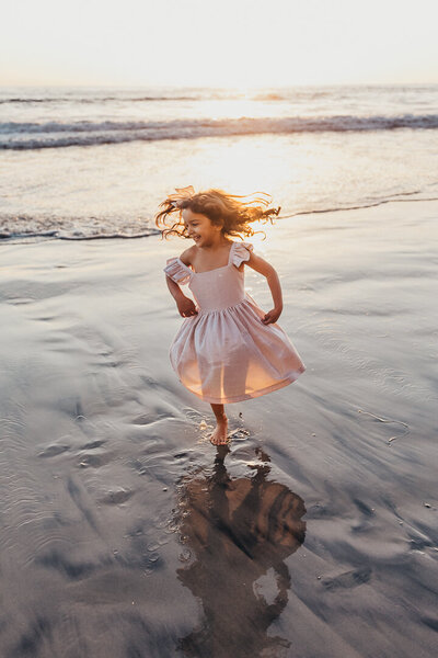 a girl in a pink dress twirls next to the ocean  on a San Diego beach