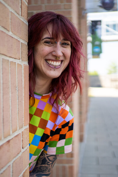 Sarah peeks out from behind a brick wall, wearing a rainbow checkered shirt