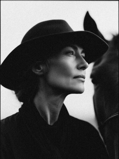 A woman in a dark hat and coat stands beside a dark horse, both looking toward the camera in a black-and-white photo—a timeless image reflecting Sweet Pea Ink Creative’s eye for brand design.