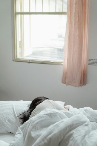 A woman lying in bed with a blanket, bathed in soft natural light, creating a serene and restful scene.