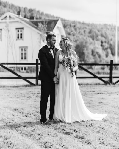A bride and groom standing in front of a wedding barn, looking at each other with the bride holding a bouquet