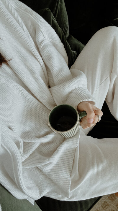 woman wearing white sitting on a green velvet chair holding a green and white tea mug