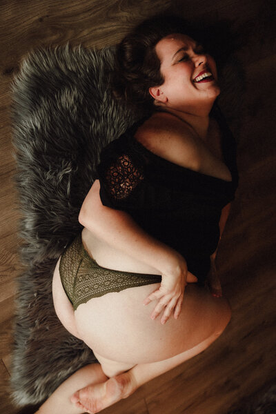 Woman in green lace underwear and black top laughs joyfully while reclining on a faux fur rug during a boudoir session in Tampa, Florida.