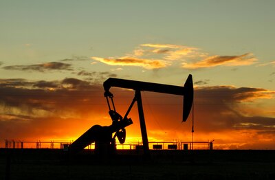 A silhouette of an oil pump jack set against a dramatic sunset with vibrant orange and yellow hues filling the sky.