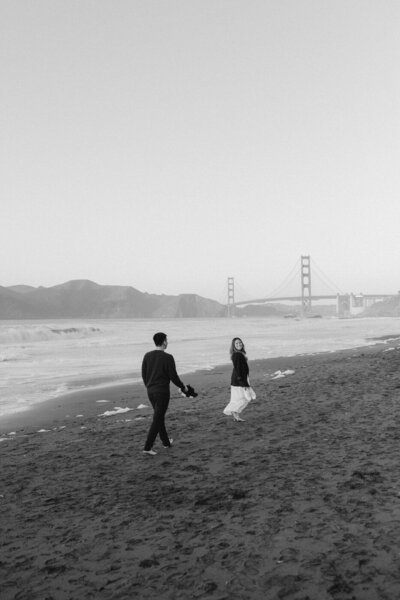 black and white classic shot of couple walking on baker beach with the golden gate bridge and san francisco in the background