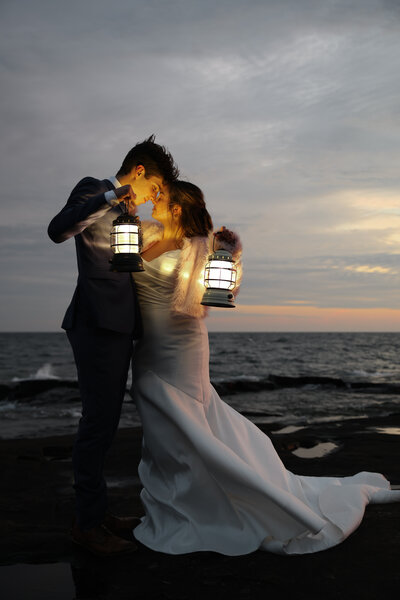 Peaceful elopement scene with a couple holding lanterns by the water as the sun dips below the horizon.