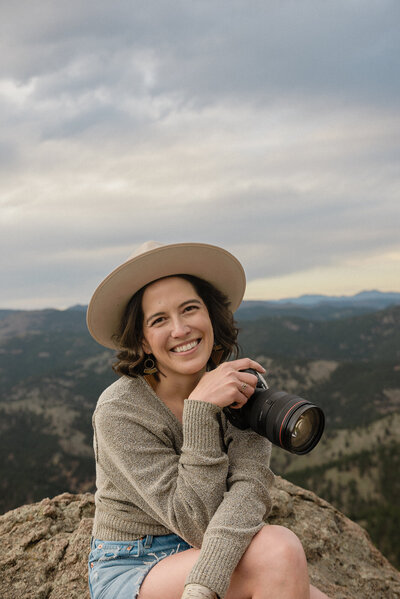 Southwest elopement photographer sits on rock overlooking the colorado mountains and poses with her camera,.