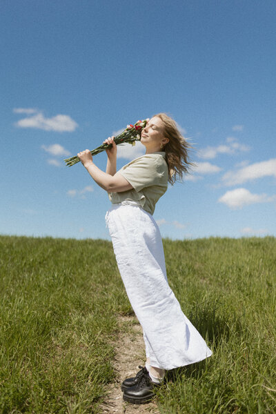A young woman holding flowers while walking along a grassy path, captured in soft natural light.