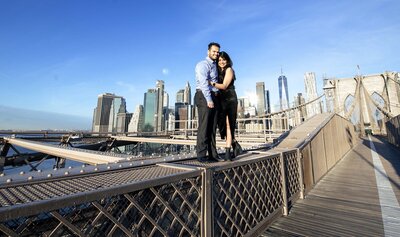 Engaged Indian couple standing on Brooklyn Bridge rails with the city in the background.