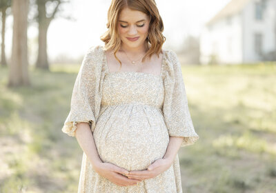 Red-haired pregnant woman in a cream lace gown holding her bump outdoors during golden hour.